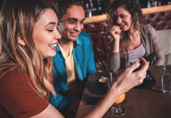 Group of cheerful young friends looking at smart phone while sitting in cafe.