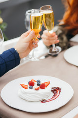 beautiful small strawberry pavlova meringue cakes with cream and fresh berries on white plate. young couple clink glasses together sitting at table in restaurant, celebrating anniversary