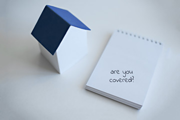 A layout of a house with a classic blue roof stands on a white surface next to the notebook with text Are you covered. Real estate insurance concept.