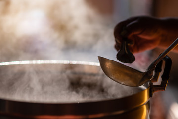The chef in the restaurant is cooking while using the dipper in a large pot. The water is boiling and the mass of steam reflected in the morning light. - obrazy, fototapety, plakaty