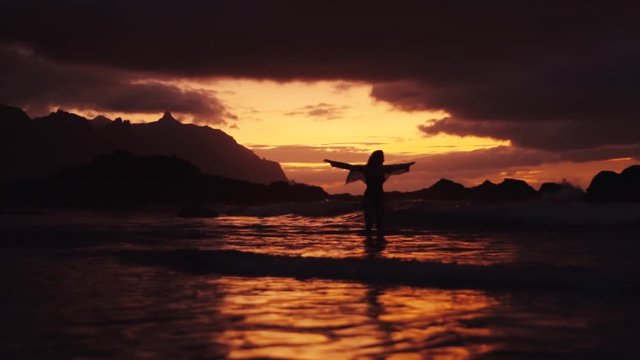 Silhouette of slim young woman walking in the sea above the red sunset sky catching waves relaxing enjoying dramatic travel evening nature rocks stones alone slow motion