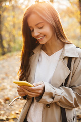 Image of young woman using smartphone while walking in autumn park