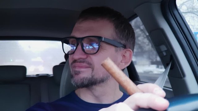 White bearded man behind steering wheel of passenger car holds cigar, smiles, nod to music.