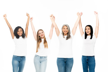 happy multicultural women in denim jeans standing with hands above head isolated on white