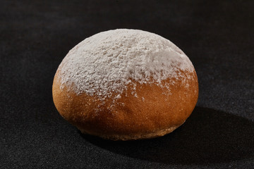 Fresh, tasty baked bun sprinkled with flour or powdered sugar against black background with copy space. Rural cuisine or bakery. Close-up