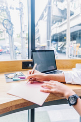 Close up of woman's hand writing in notebook placed on wooden desktop with various items and street view