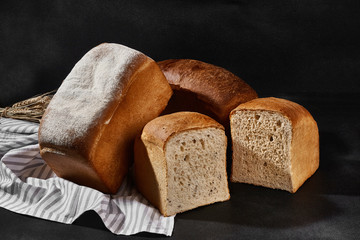 Variety of delicious baked bread, whole and cut in halves, sprinkled with flour and bunch of wheat are on a kitchen towel. Black background. Close-up