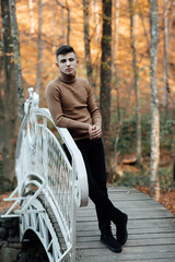 Fototapeta na wymiar Teen guy walks through the woods in autumn. Pensive and funny teenager. Cute spectacled smiling happy teen boy, looking at camera. Kids outdoor portrait. The guy rests on the railing of the bridge