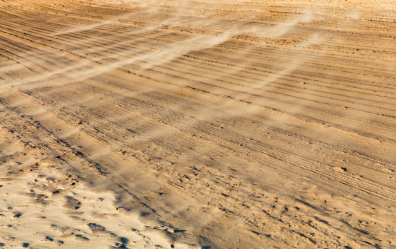 Sand background, sandy waves on windy day on the beach or in desert, natural background. Strong wind on the beach, sand storm. Sand is flying in the face. Tire tracks on a sandy beach.