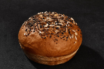 Fresh, palatable baked bun sprinkled with sesame and sunflower seeds against black background with copy space. Rural cuisine or bakery. Close-up