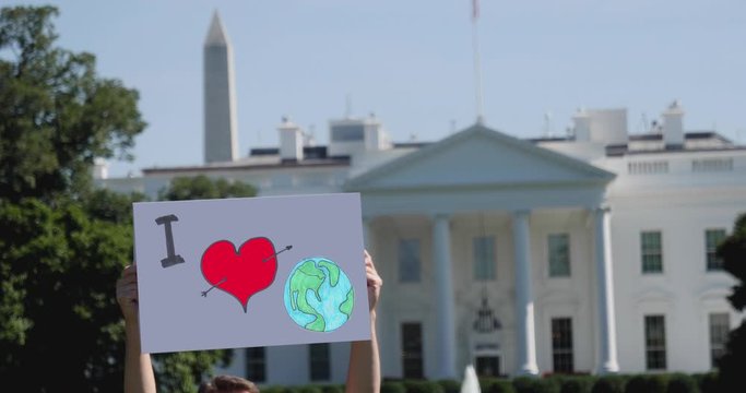 A man holds a I LOVE EARTH protest sign in front of the White House on a sunny summer day.  	