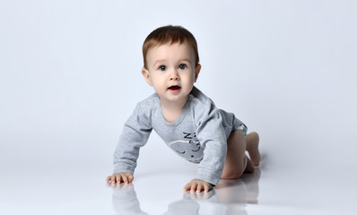 Little baby boy toddler in grey casual jumpsuit and barefoot crawling on floor, smiling and looking up over white wall background