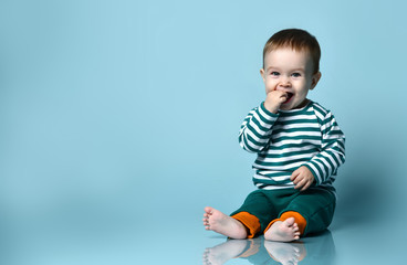 Little baby boy in stylish casual clothing barefoot sitting on floor and smiling over blue wall...
