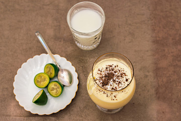 A gourmet bowl with a cottage cheese dessert sprinkled with chocolate is on the table, next to a saucer with green feijoa and a glass with a milkshake