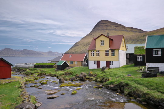 Horizontal scenery image of Faroese landscape with idyllic village Gjogv, most northern village on the island of Eysturoy in foreground and high mountains in background covered with clouds