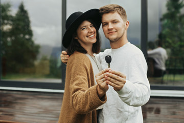 Home and family concept. Stylish hipster couple holding together key from their new home on background of terrace and big windows outdoors. Happy young family of homeowners, purchasing real estate