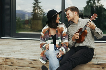 Hipster man playing on ukulele for his stylish woman, relaxing on wooden porch  of modern cabin with big windows in mountains. Happy young family travelers smiling and having fun