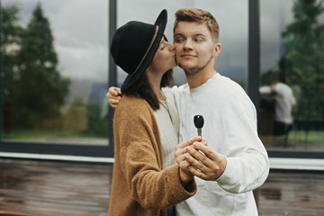 Stylish hipster couple holding together key from their new home on background of terrace and big windows outdoors. Happy young family in modern outfits celebrating buying first property