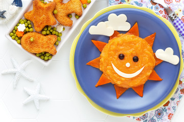 Fun food for kids - cute smiling sun face made of mashed potato and pumpkin puree, decorated with cheese and served with crispy fish nuggets and green peas