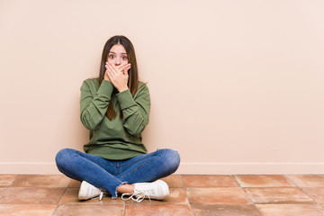 Young caucasian woman sitting on the floor isolated shocked covering mouth with hands.