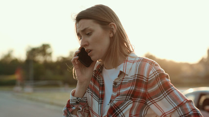 Disappointed young woman talking on the phone standing in the street at road accident. Mixed-up girl in shirt asking for help calling for the police. Car crash.