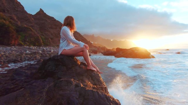Side view of caucasian woman in white shirt sitting on rock and watching at big waves crashing agains the stone and wery exited relaxing enjoying nature sunset summer evening travel slow motion