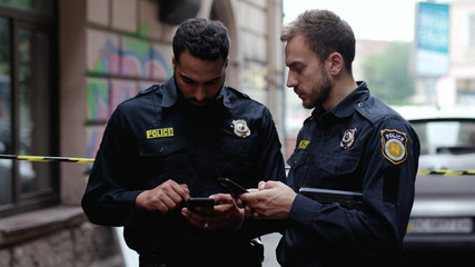 Police at raid. Close-up two handsome policemen searching on smartphones tracking a criminal online. Friendly colleagues cops at police investigation area in the city street.