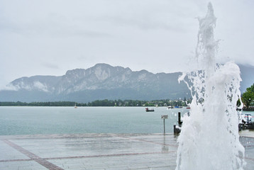 Mondsee, Austria - Fountain on the promenade by Lake Mondsee, a white stream of water on a site laid out of stone tiles, in the background water, mountains and trees.