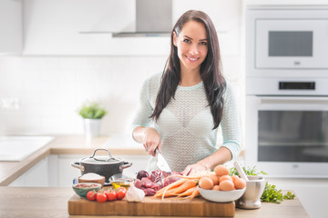 Cheerful young woman is happy to prepare lunch in her kitchen. She prepares food from meat and vegetables and other healthy ingredients