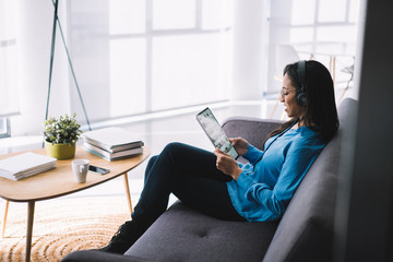 Black woman in headphones sitting with tablet on sofa and listening music