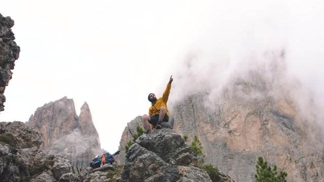 Young man hiker sitting on stone mountain with map pointing forward, with cloudy sky and fog. Yellow jacket, backpack, black beard and beanie. Traveling dolomites, Italy.