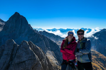 A couple on the top of Grosse Gamswiesenspitze in Lienz Dolomites, Austria. Sharp, dangerous slopes. Massive Alpine mountains. They are happy and tired after their hike. Clouds in the valley. Freedom