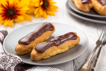 Tasty eclairs on a plate on a white wooden table. Served with flowers. Rectangular gray plate. Traditional french eclairs with chocolate. Home made cake eclairs. Traditional eclairs, profitroles class