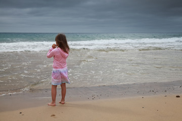 Caucasian baby girl in pink stands on the sea coast,