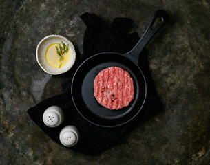 Raw Ground beef meat Burger steak cutlet with seasonings served on metal frying pan over the kitchen towel with the bowl of olive oil and salt and pepper shakers. Rustic metal background. Top view