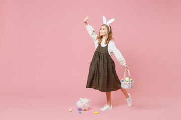 Little pretty blonde kid girl 11-12 years old in light spring dress hold fluffy white bunny rabbit, wicker basket with eggs isolated on pastel pink background. Childhood lifestyle Happy Easter concept