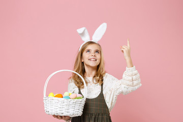 Little pretty blonde kid girl 11-12 years old in spring dress, bunny rabbit ears hold in hand wicker basket colorful eggs isolated on pastel pink background. Childhood lifestyle Happy Easter concept.