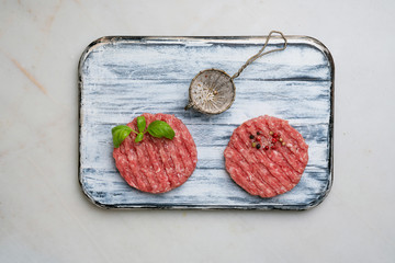 Raw Ground beef meat Burger steak cutlets with seasonings and herbs served on rustic wooden board with metal strainer. White marble background. Top view