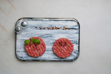 Raw Ground beef meat Burger steak cutlets with seasonings and herbs served on rustic wooden board with salt and pepper shakers over white marble background. Top view