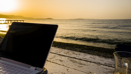 Freelance working with laptop on wooden table on beach at sunset. Woman sitting on chair and using computer near blue sea shore at sunny day. Digital nomadism towards yellow sky & horizon