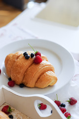 Croissant decorated with fresh raspberries and blueberries on a white plate and a wooden white tray. Tasty breakfast. Bakery products