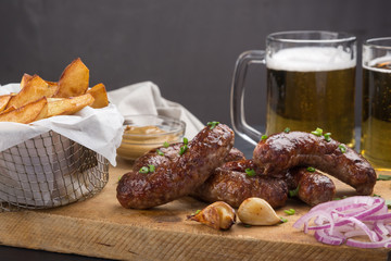Grilled sausages from a BBQ served with a mustard, fried potatoes, garlic, herbs, beer on a wooden board all served on a black wooden table. Delicious german sausages Perfect for Octoberfest.