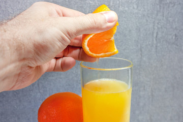 Male hand squeezes juice from a half orange into a glass cup on a gray background. On a white table lies a whole orange.