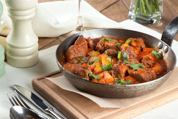 Stewed rabbit with potatoes and vegetables served in a frying pan. Served on a table with white napkin, fork, knife, wine, flowers.
