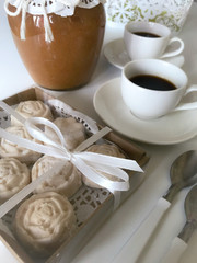 Box of coconut candies in the shape of flowers. Tied with a ribbon tied to a bow. Near cups with coffee and a can of jam.