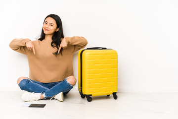 Young chinese traveler woman sittting on the floor with a suitcase isolated points down with fingers, positive feeling. © Asier