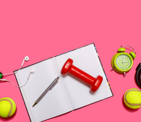 Healthy lifestyle concept with empty exercise book for text, fitness and sport. Tennis ball with dumbbells and green alarm clock isolated on pastel pink background.