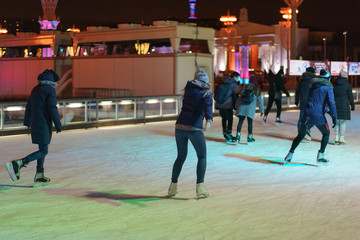 Fototapeta premium Ice skating rink in the city park. Men and women are here. Youth skating and getting fun in night time. Sportive lifestyles concepts. Backs.