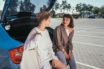 Two girls in the parking lot at the open trunk posing for the camera.