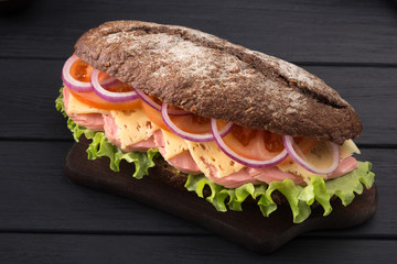 Delicious sandwich on a black wooden table. Sandwich with meat, ham, cheese, lettuce, tomatoes, onion, sauce. Close-up shot. Sandwich bread tomato, lettuce and yellow cheese. Free space for text.